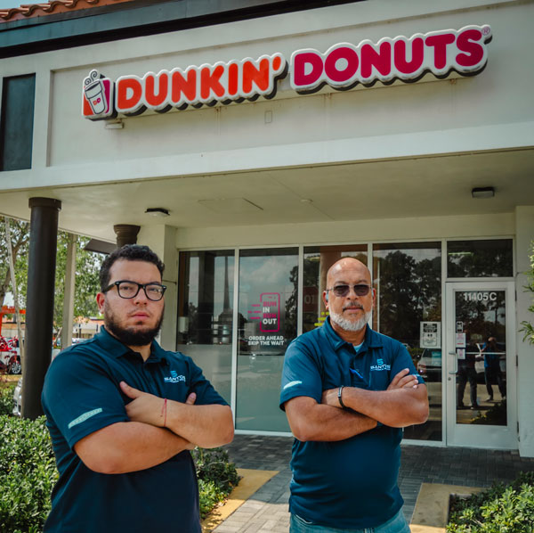 CEO and founder of Santos Technical Services, along with his son and technician, standing confidently with arms crossed in front of a Dunkin' Donuts location. Experts in commercial kitchen maintenance and repair, specializing in coffee machines, refrigeration, and ice equipment in Boca Raton, Florida.